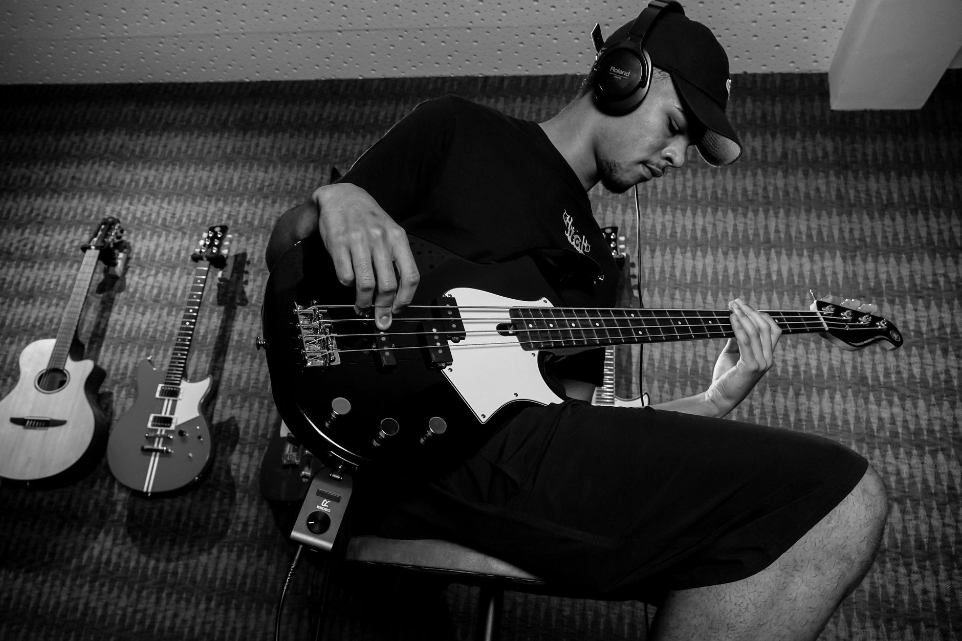 A focused musician plays bass guitar in a studio, surrounded by various guitars and headphones, captured in black and white.