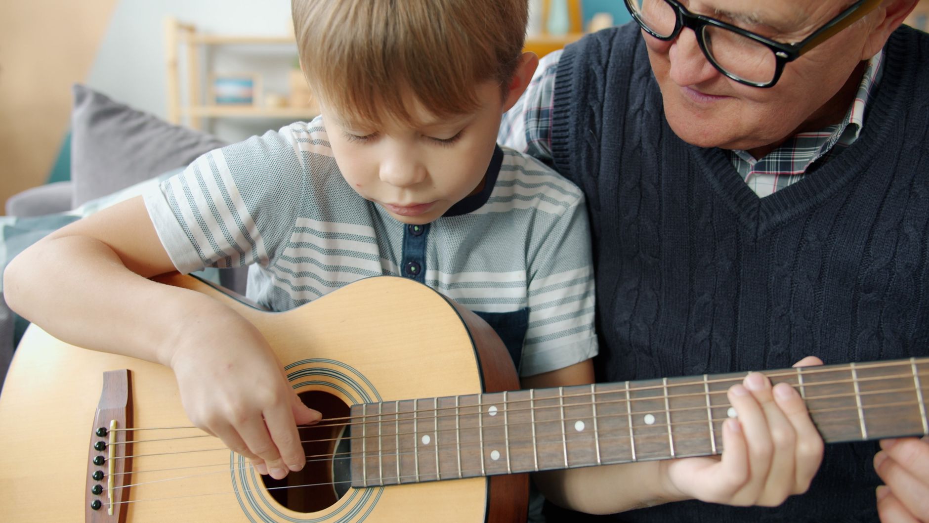 Grandfather guiding grandson on guitar playing at home, bonding moment.
