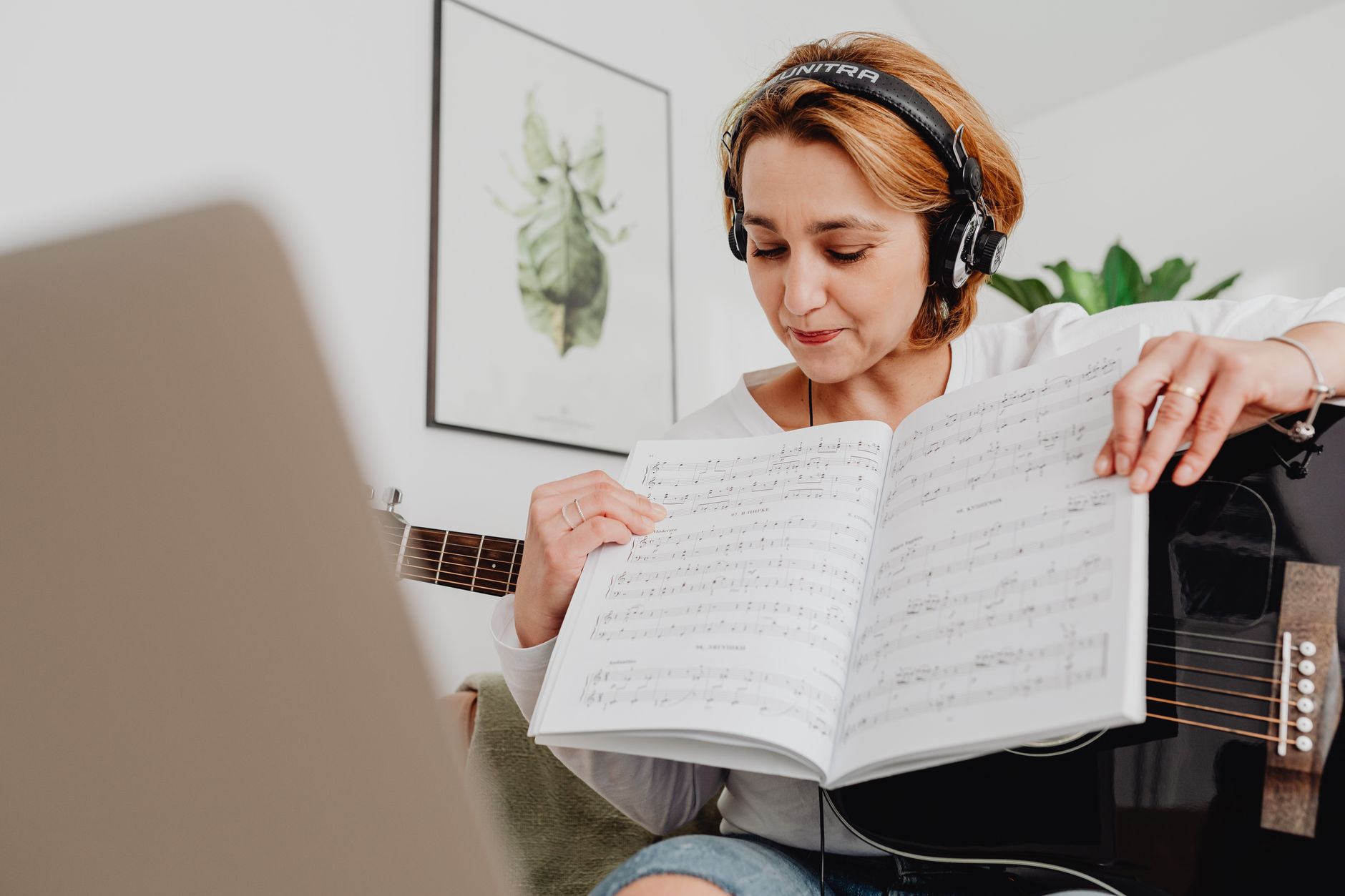 Woman practicing guitar at home using sheet music and laptop, creating a musical atmosphere.