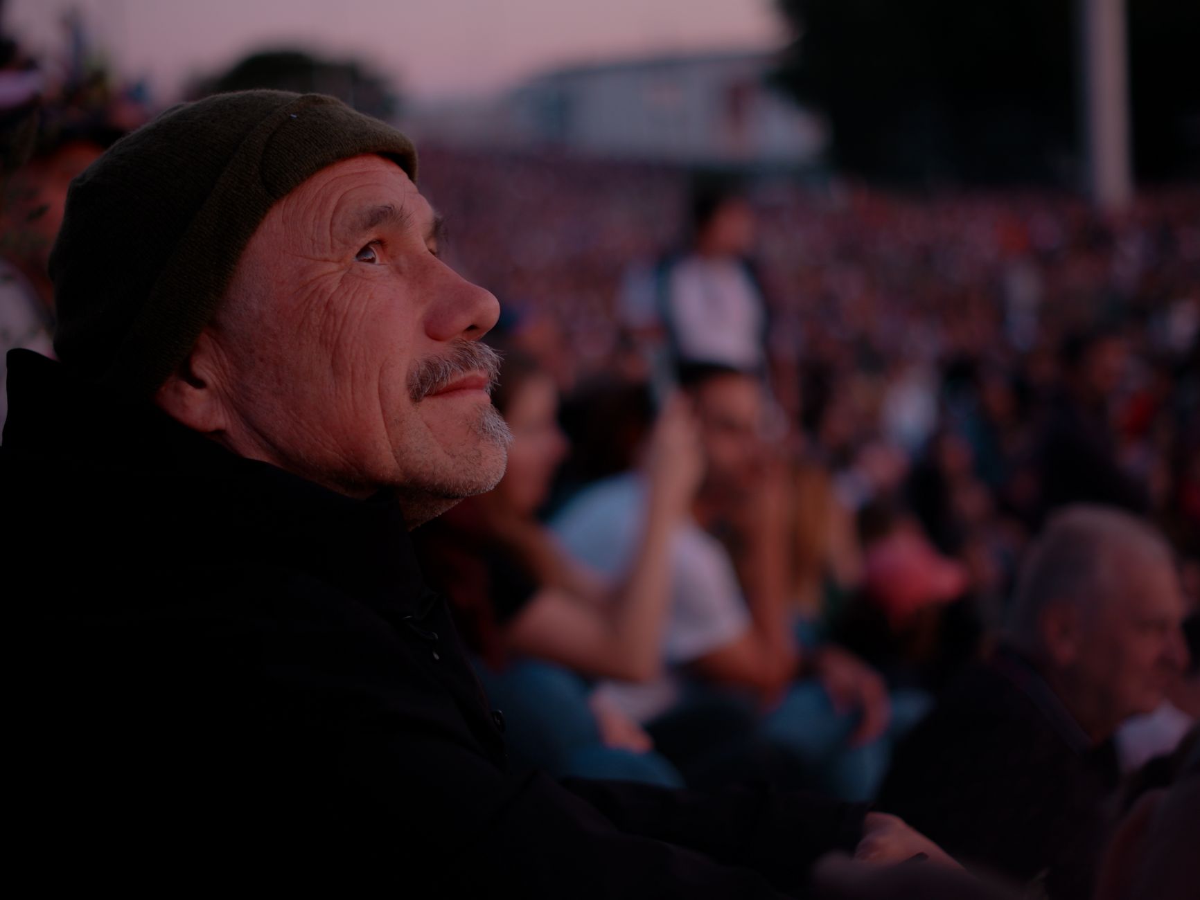 An elderly man sits among a crowd, captivated by an outdoor concert at twilight in Bratislava.