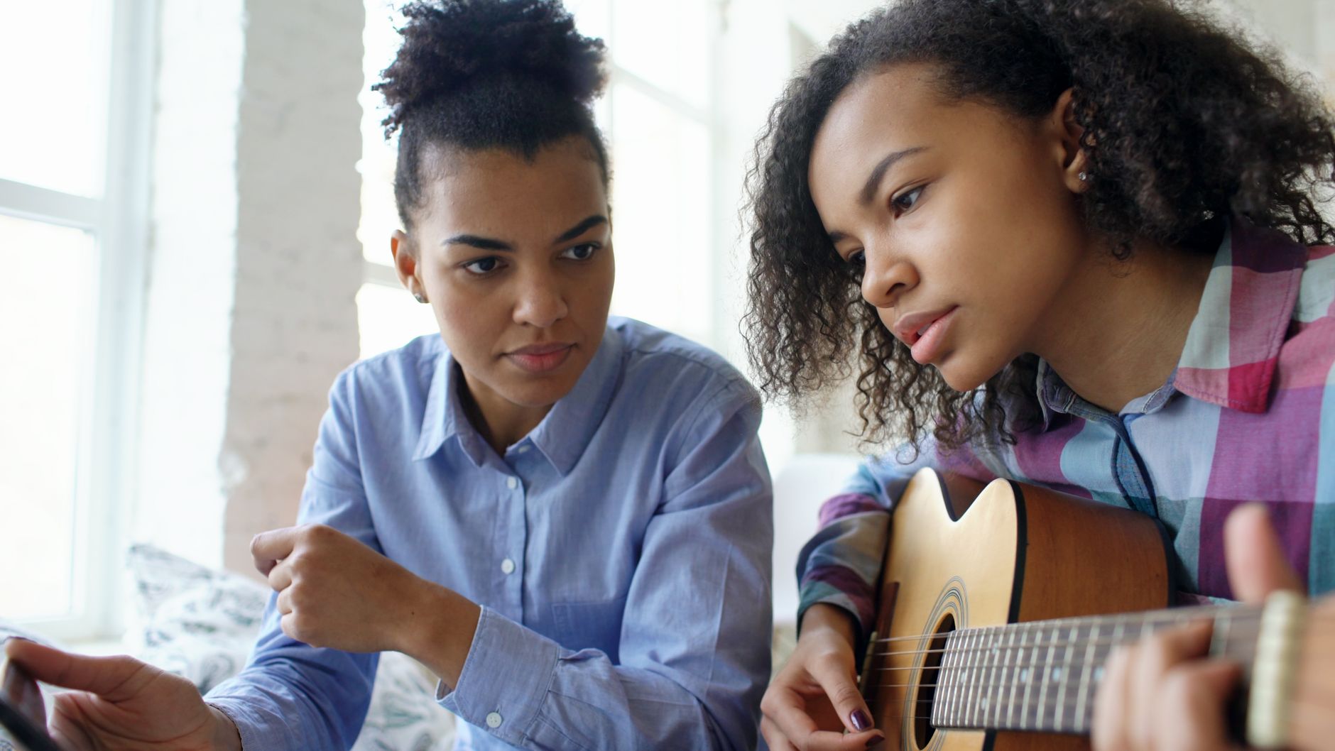 Woman teaching her daughter to play guitar indoors on a sunny day.