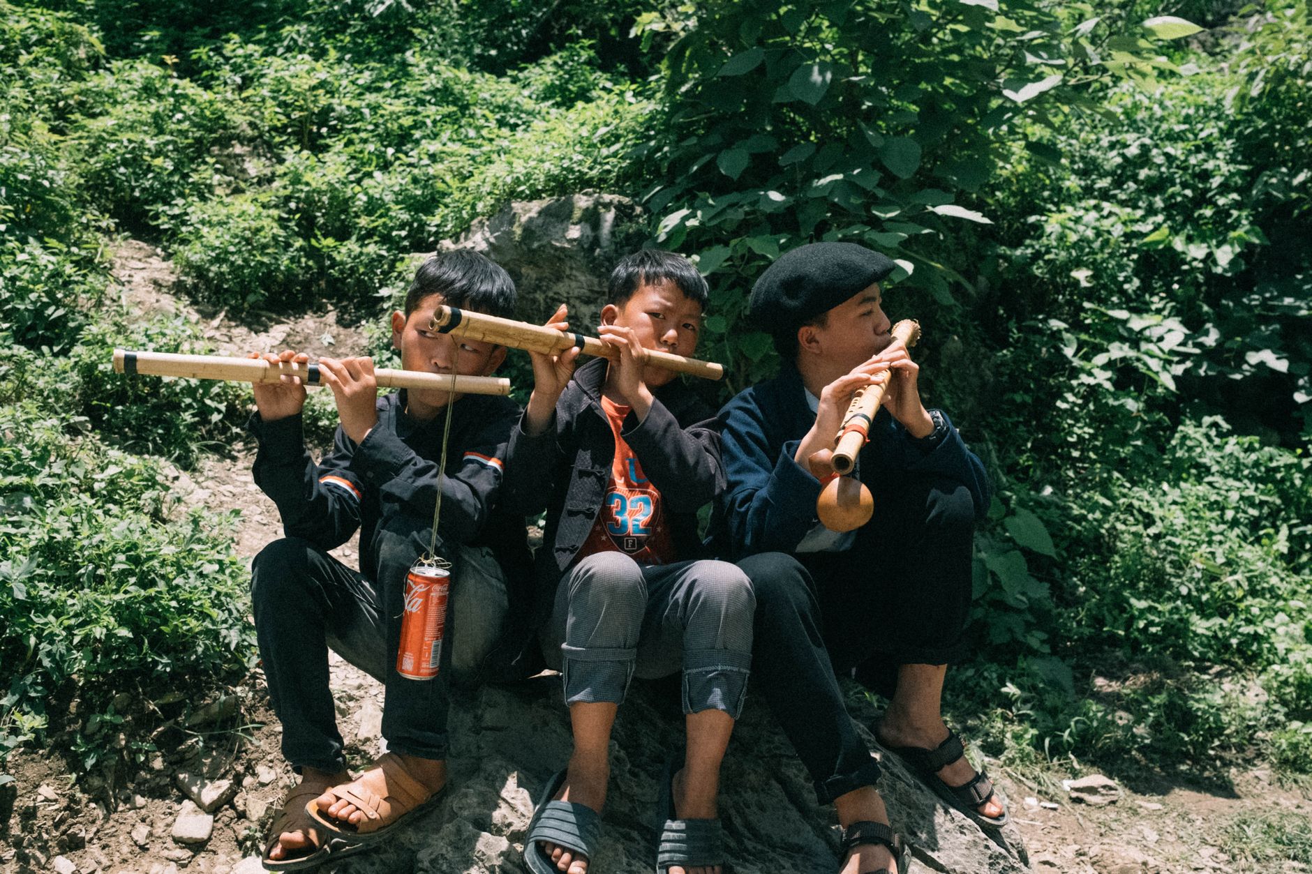 A group of children sitting outdoors playing bamboo flutes on a sunny day.