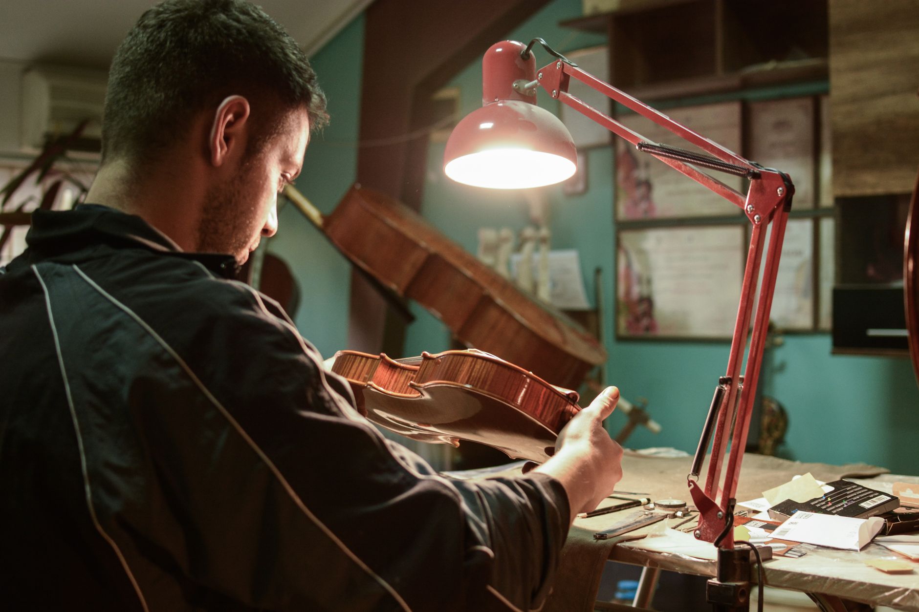 A skilled luthier carefully examines a violin in his workshop, illuminated by a desk lamp.