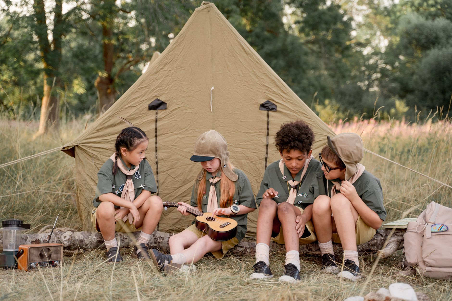 A group of children in scout uniforms sitting by a tent playing a ukulele, enjoying the outdoors.