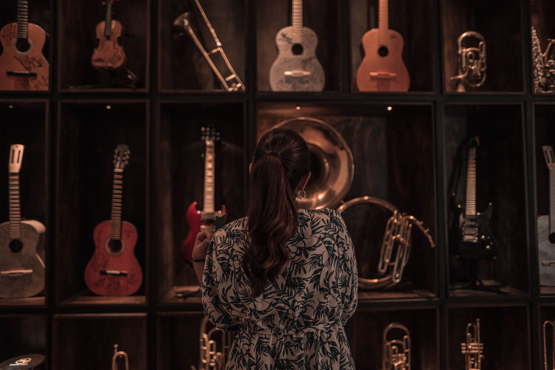 A woman observes a diverse collection of musical instruments on display, including guitars and trumpets, in a dimly lit room.