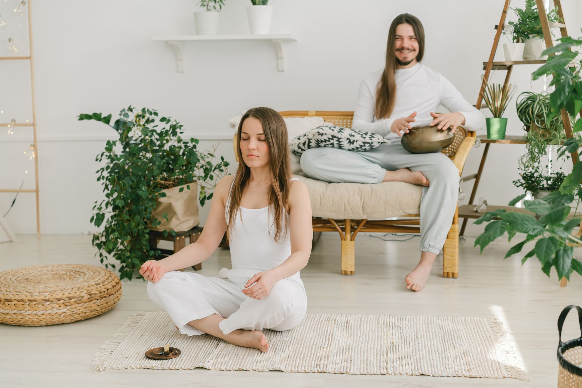 A serene indoor meditation session with a man playing a handpan and a woman meditating, surrounded by greenery.