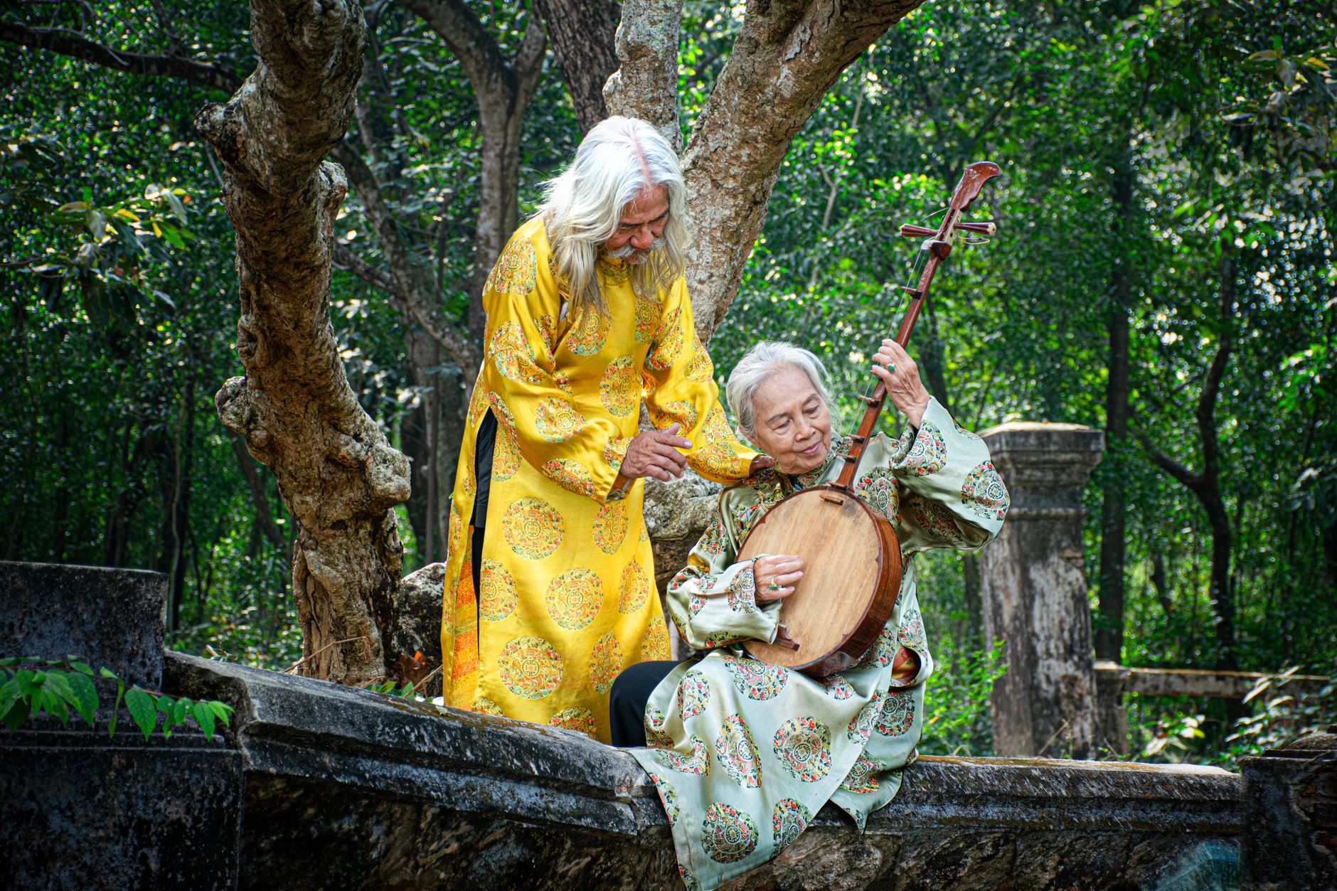 Elderly couple in traditional attire playing a Đàn Nguyệt outdoors in Bình Dương, Việt Nam.