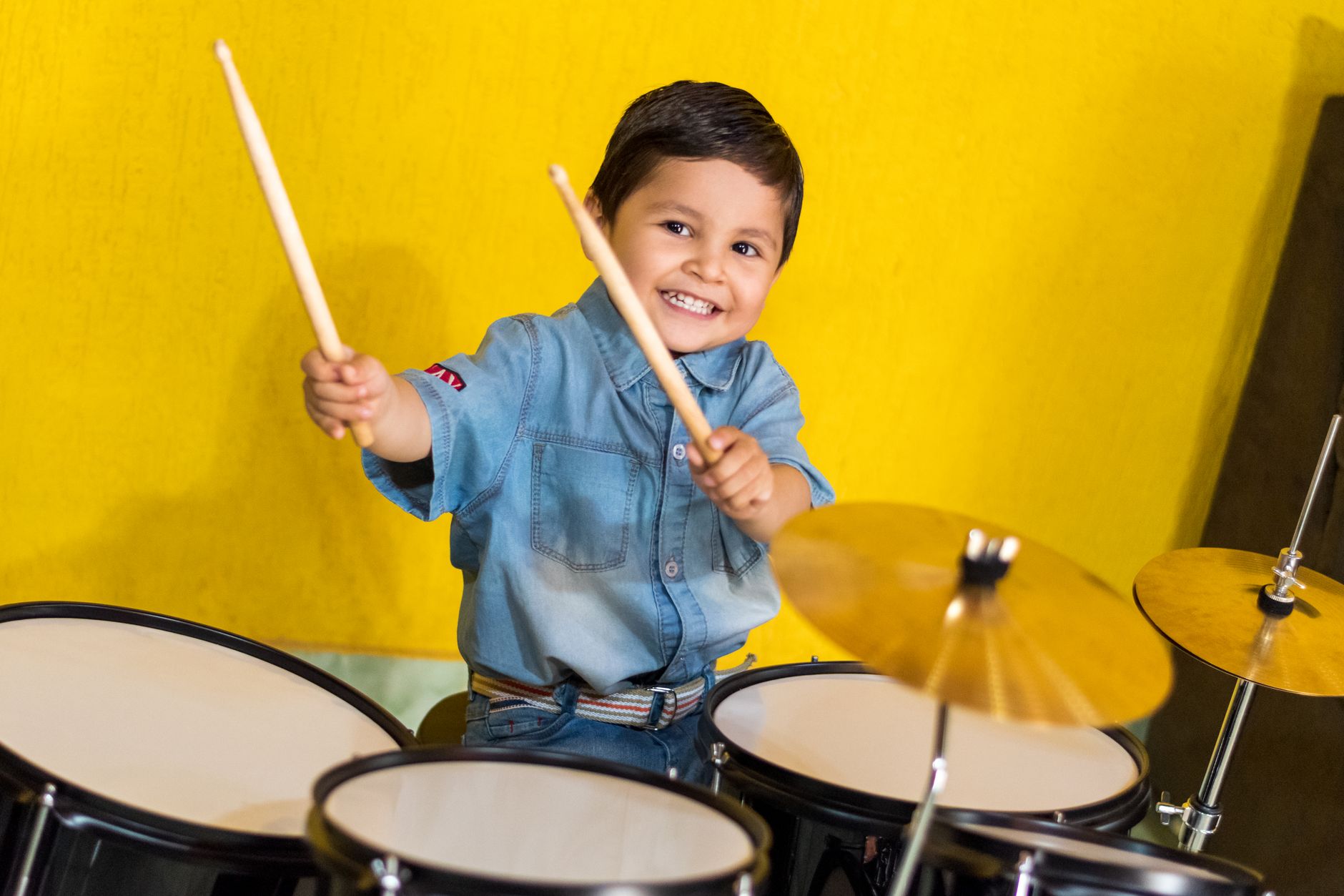 Smiling young child enthusiastically playing a drum set in a bright indoor setting.