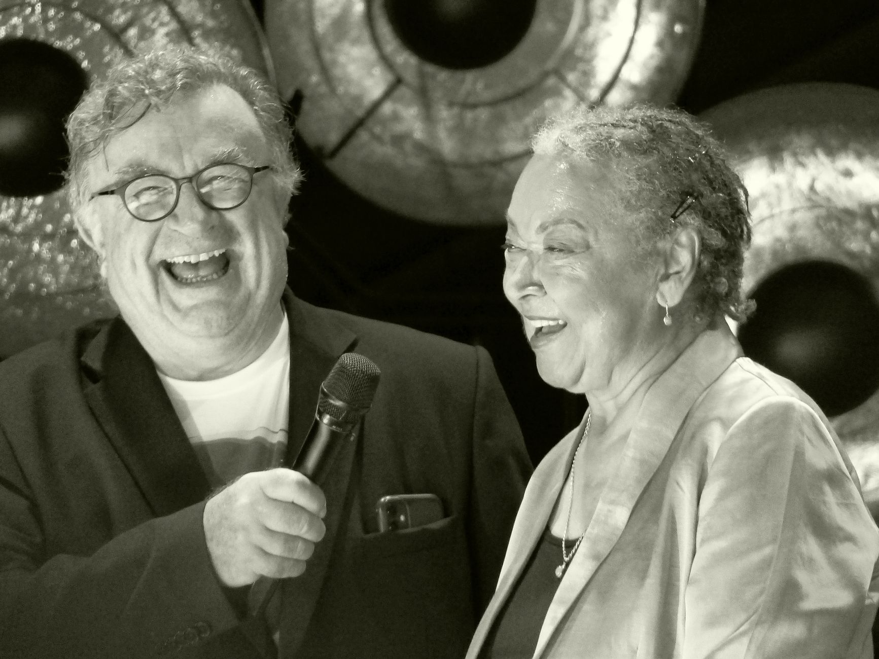 Elderly man and woman sharing laughter and joy on stage holding a microphone.
