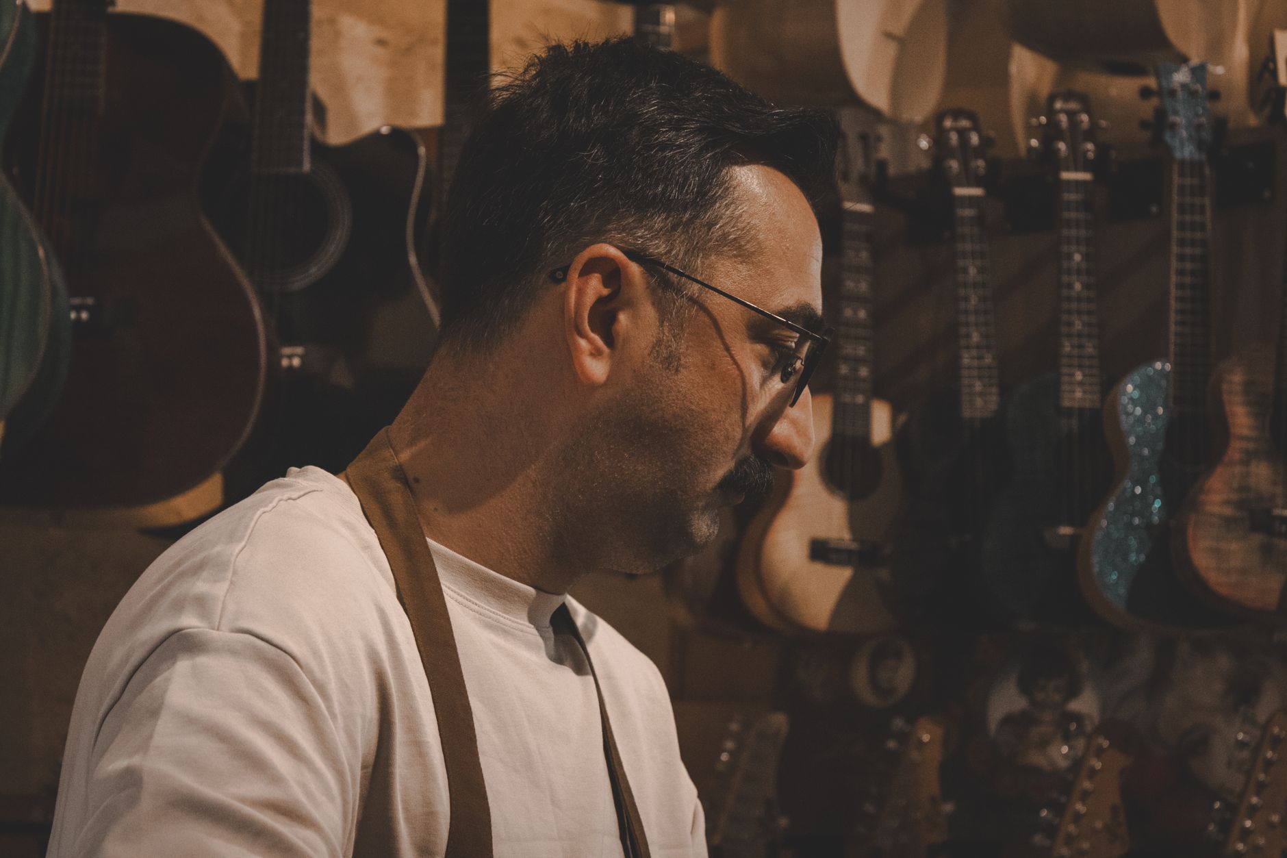 A luthier works intently surrounded by guitars and ukuleles in an artisan workshop.