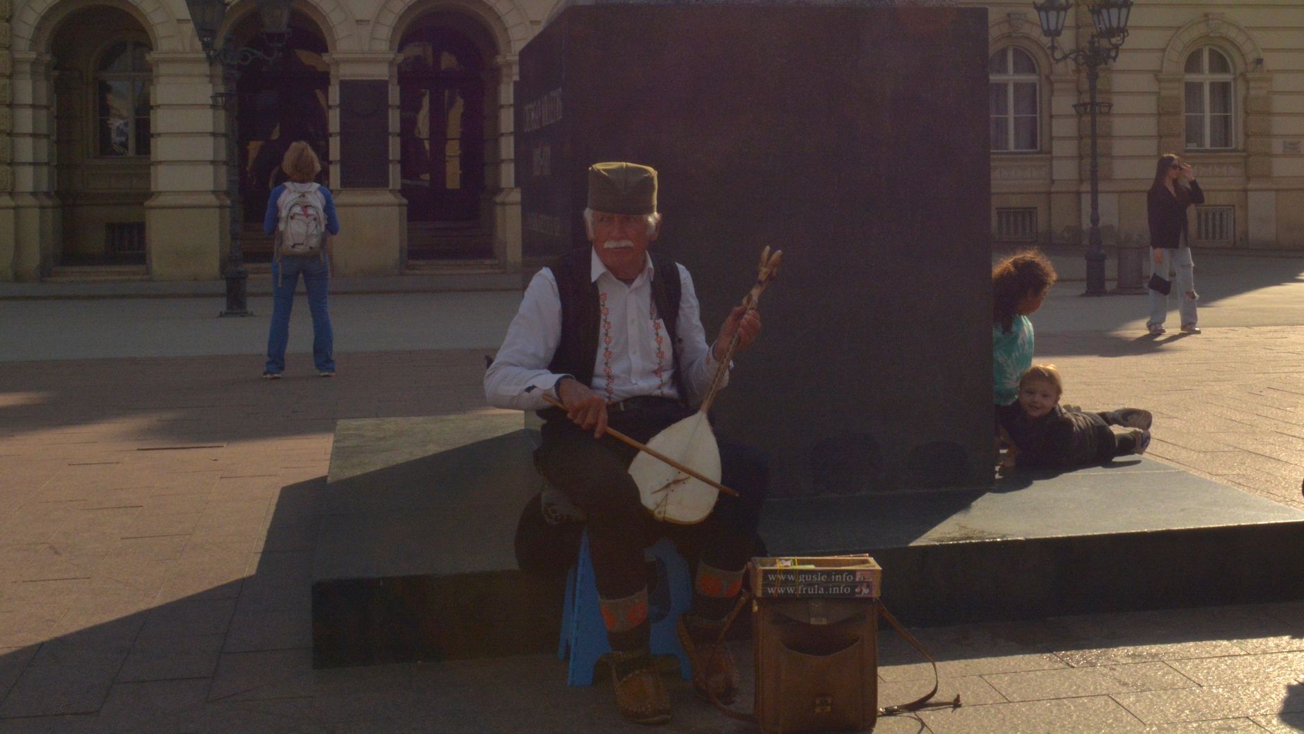 Elderly musician performs outdoors playing a gusle in an urban setting.