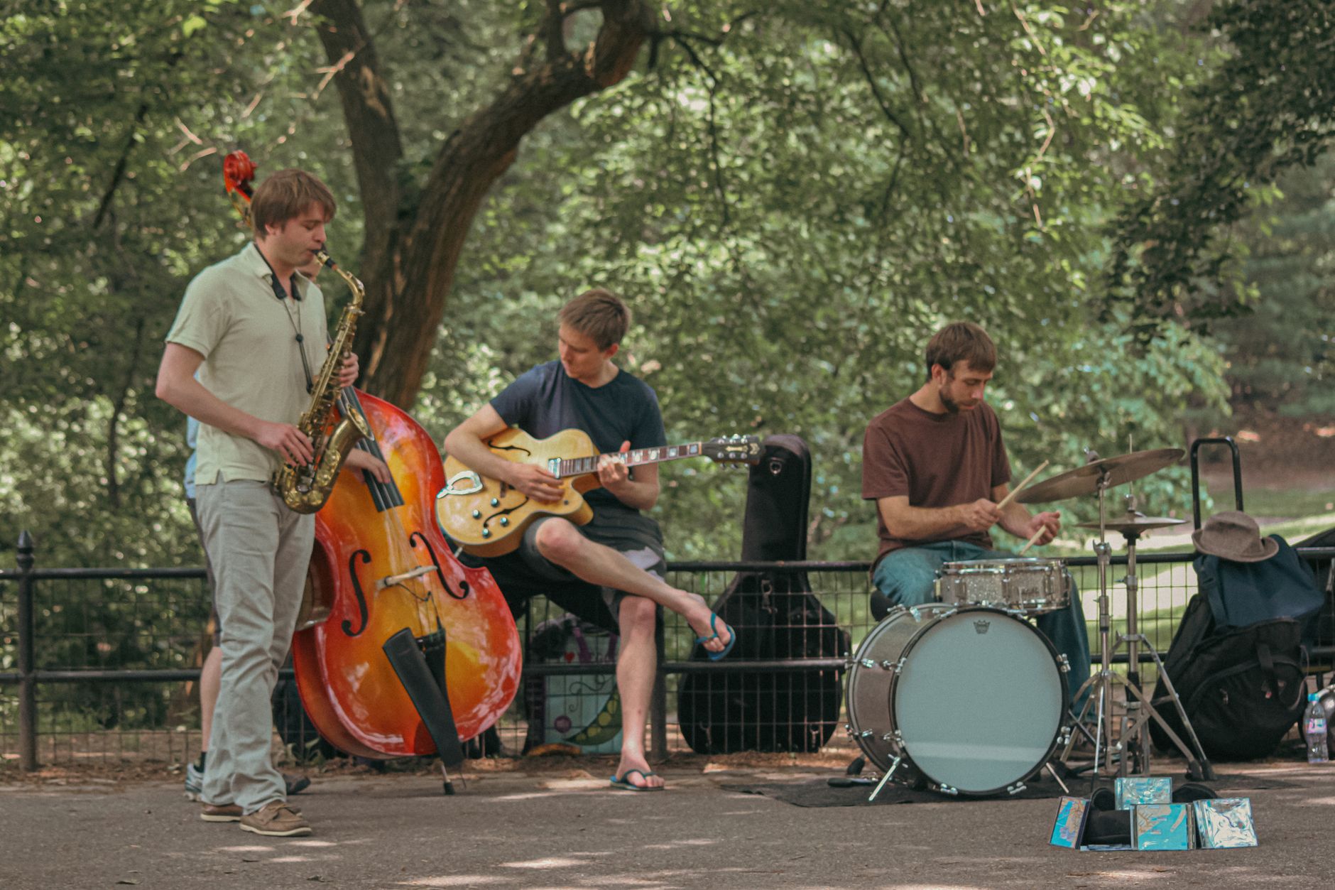 Three musicians perform in a New York City park, playing saxophone, guitar, and drums.