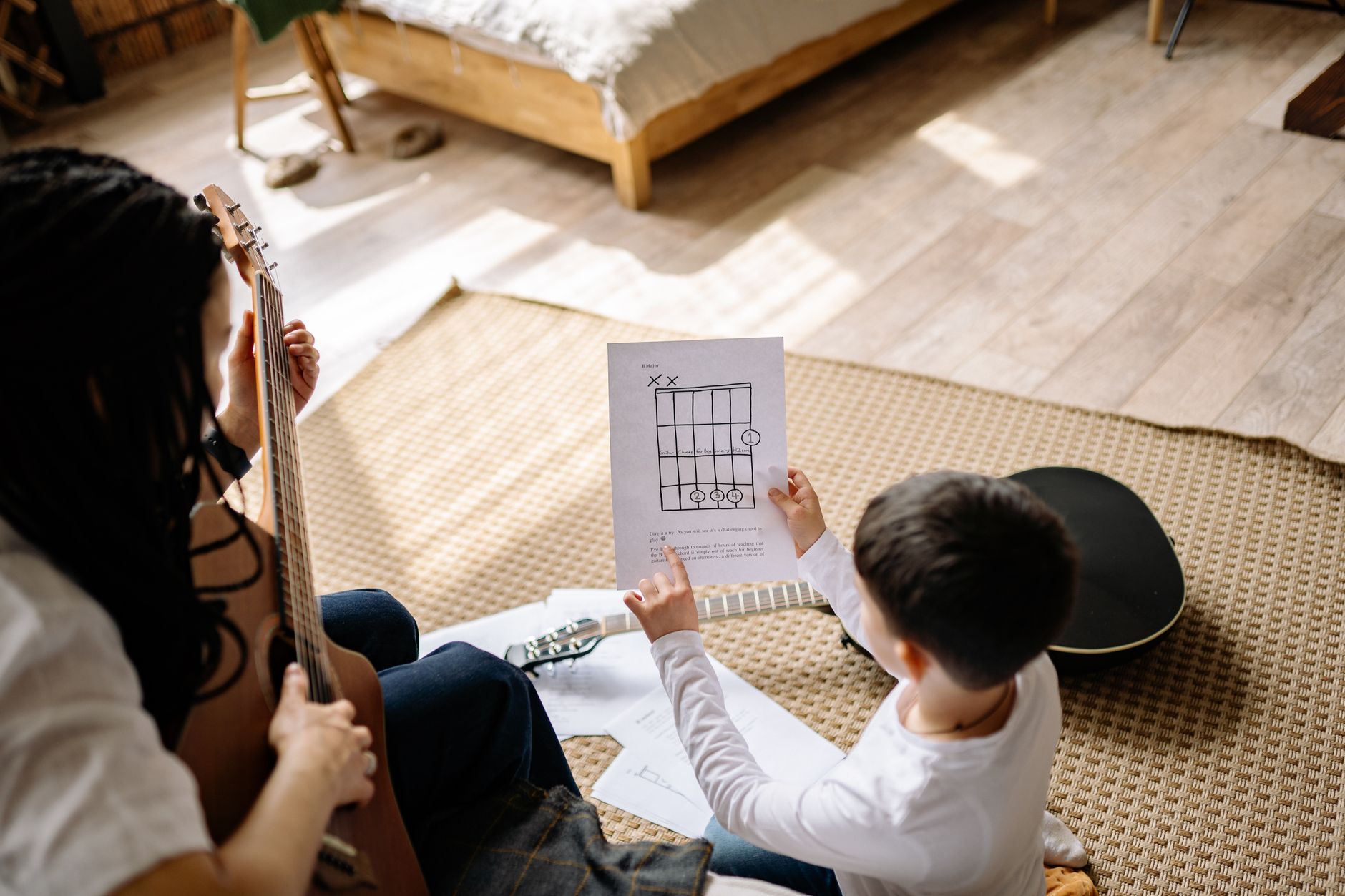 A boy learns guitar chords at home with his mother using sheet music.