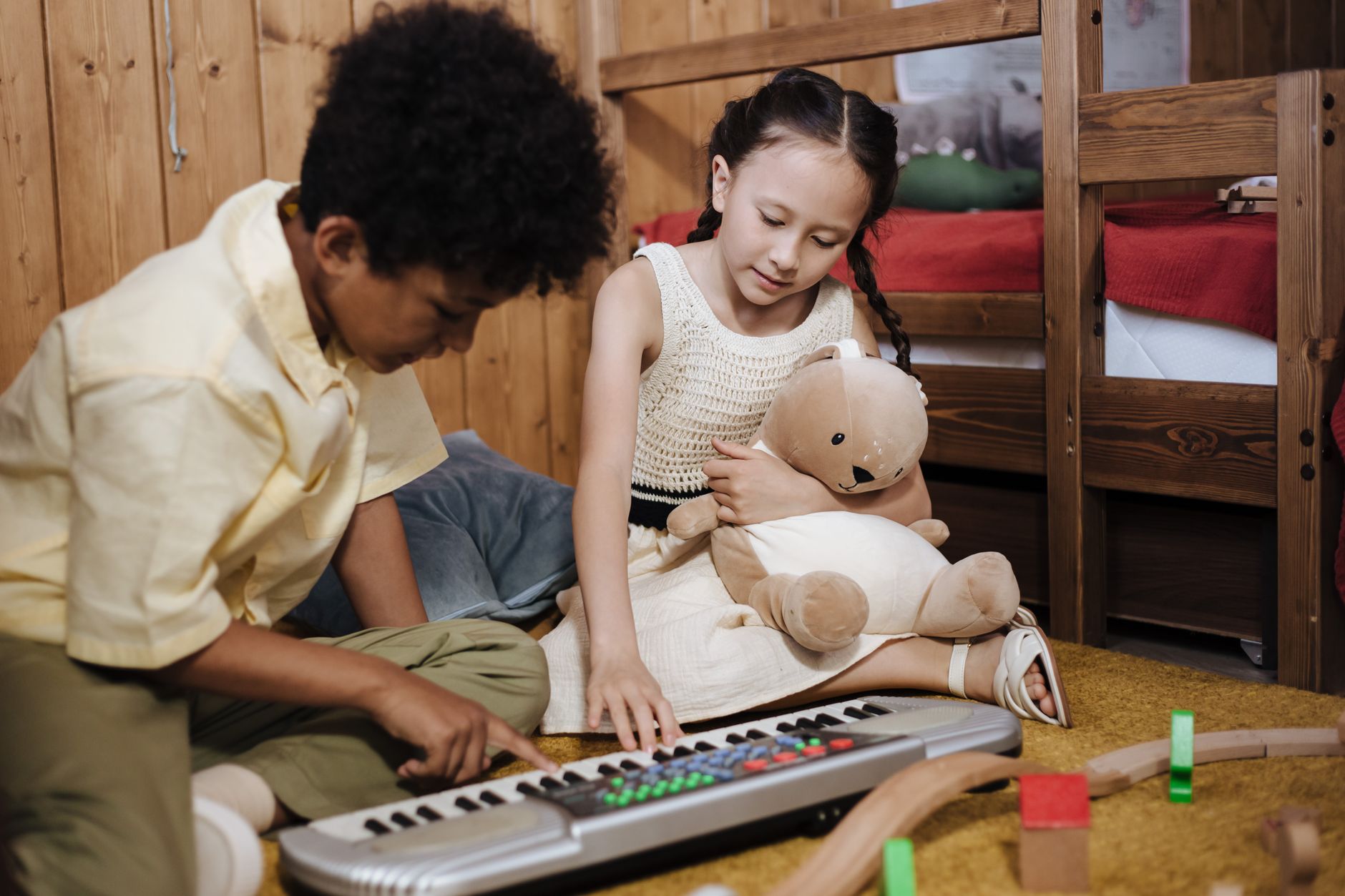 Two children playing a synthesizer in a cozy, wooden interior setting with a teddy bear.
