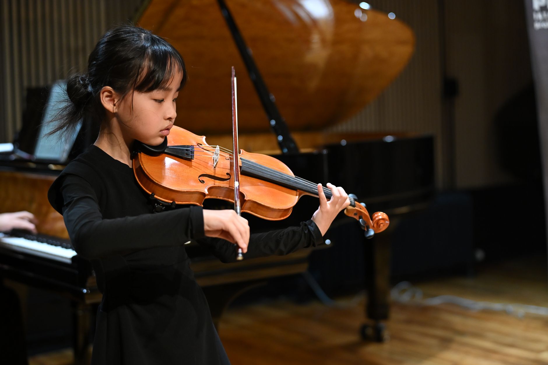A young girl passionately playing the violin on stage, with a grand piano in the background.