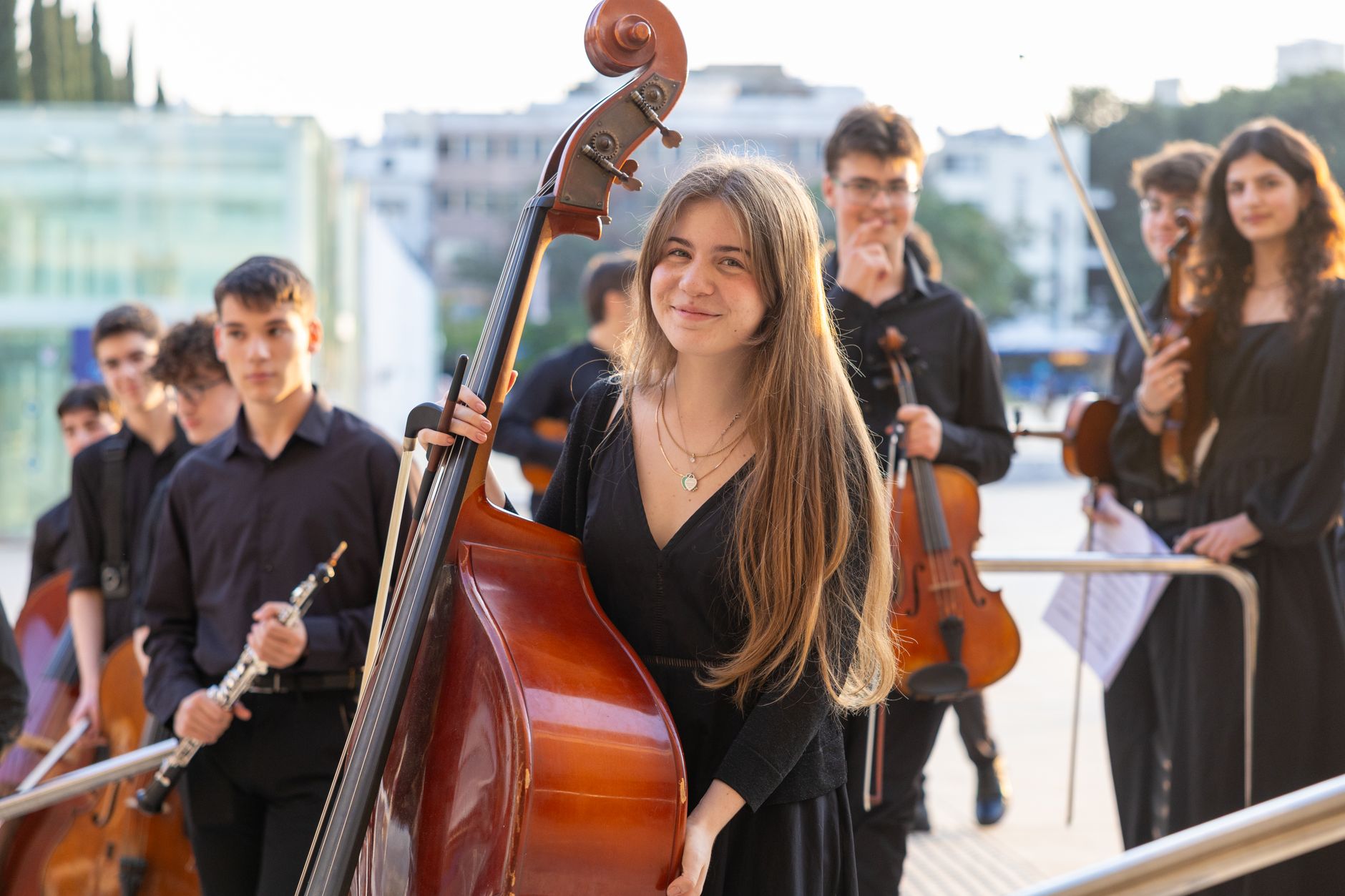 A group of young musicians in an outdoor setting preparing for a performance, featuring a young woman with a double bass.
