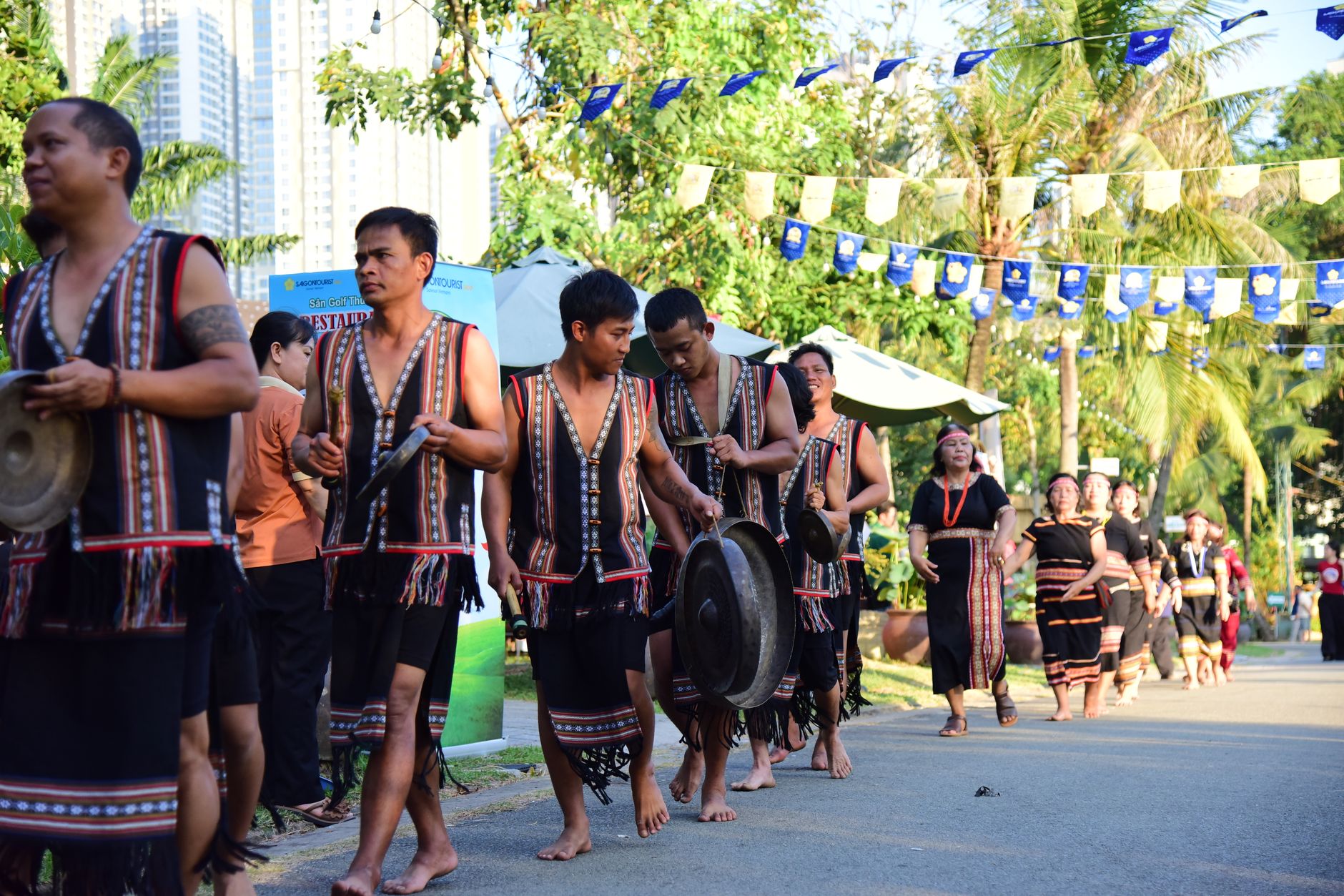 Men and women in traditional attire performing with gongs during a cultural parade outdoors.