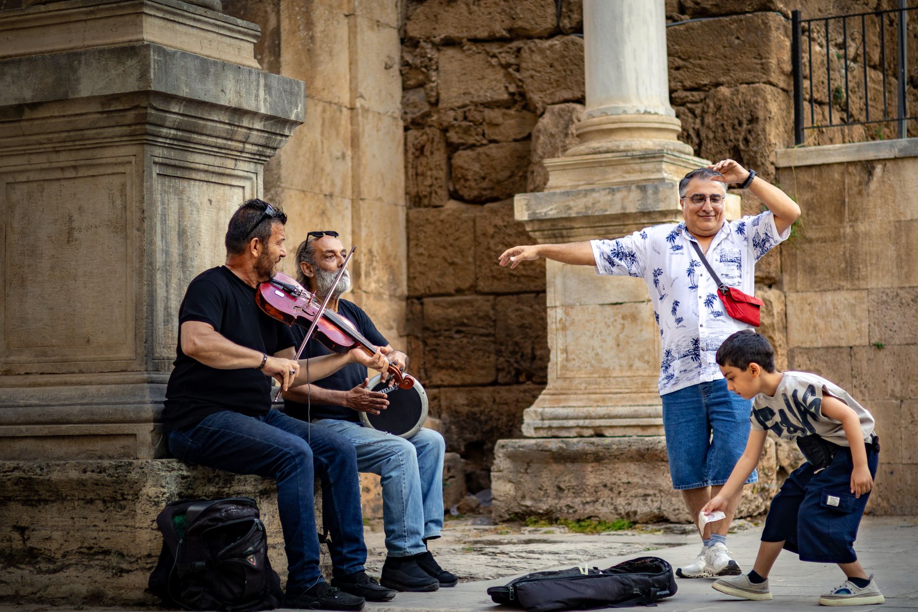 Two street musicians play with a violin and drum, while passersby enjoy and a child shows interest.